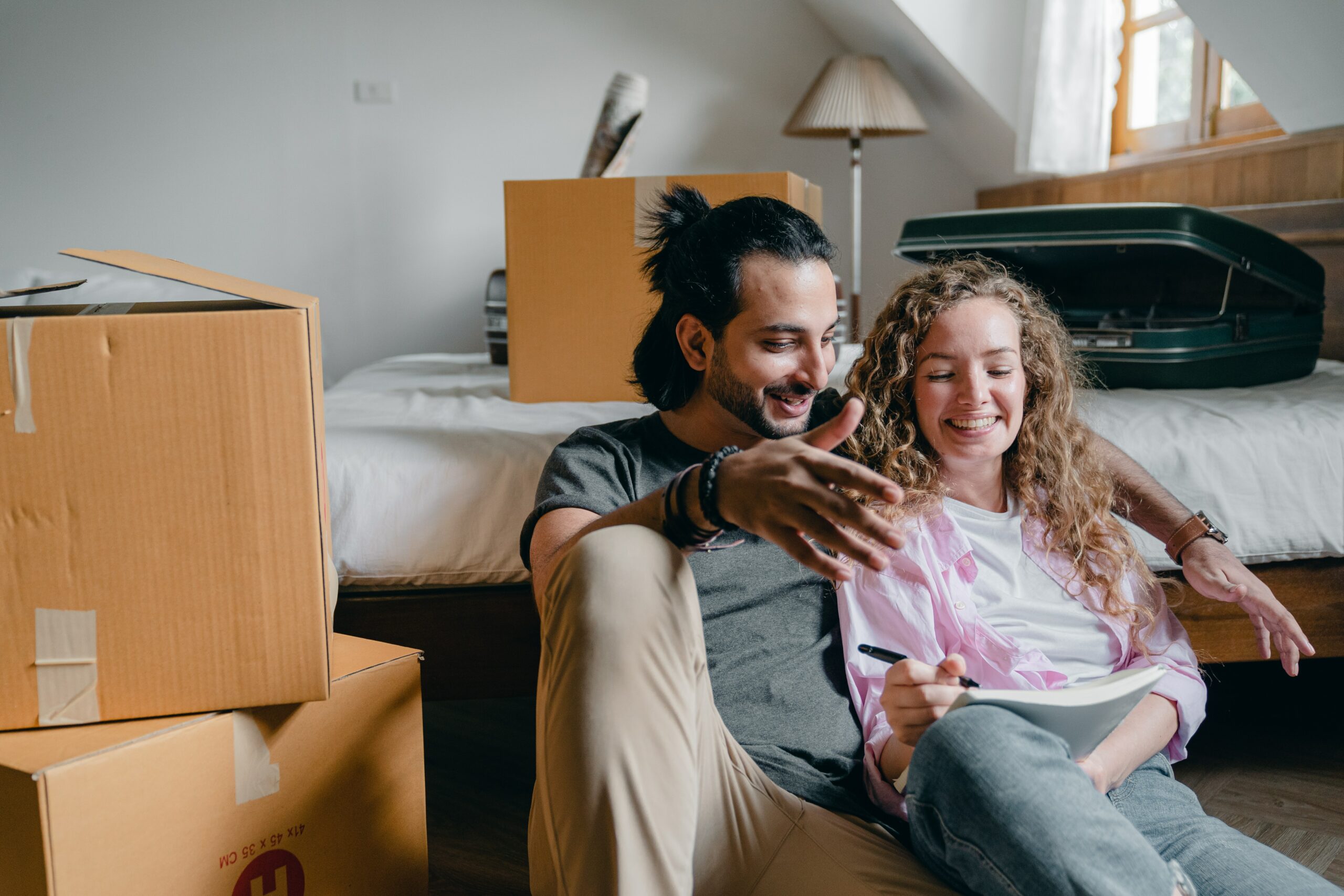 Happy couple sitting in bedroom next to cardboard moving boxes. 