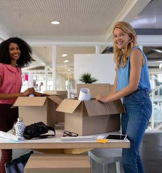 Two women pack boxes in a modern office setting.